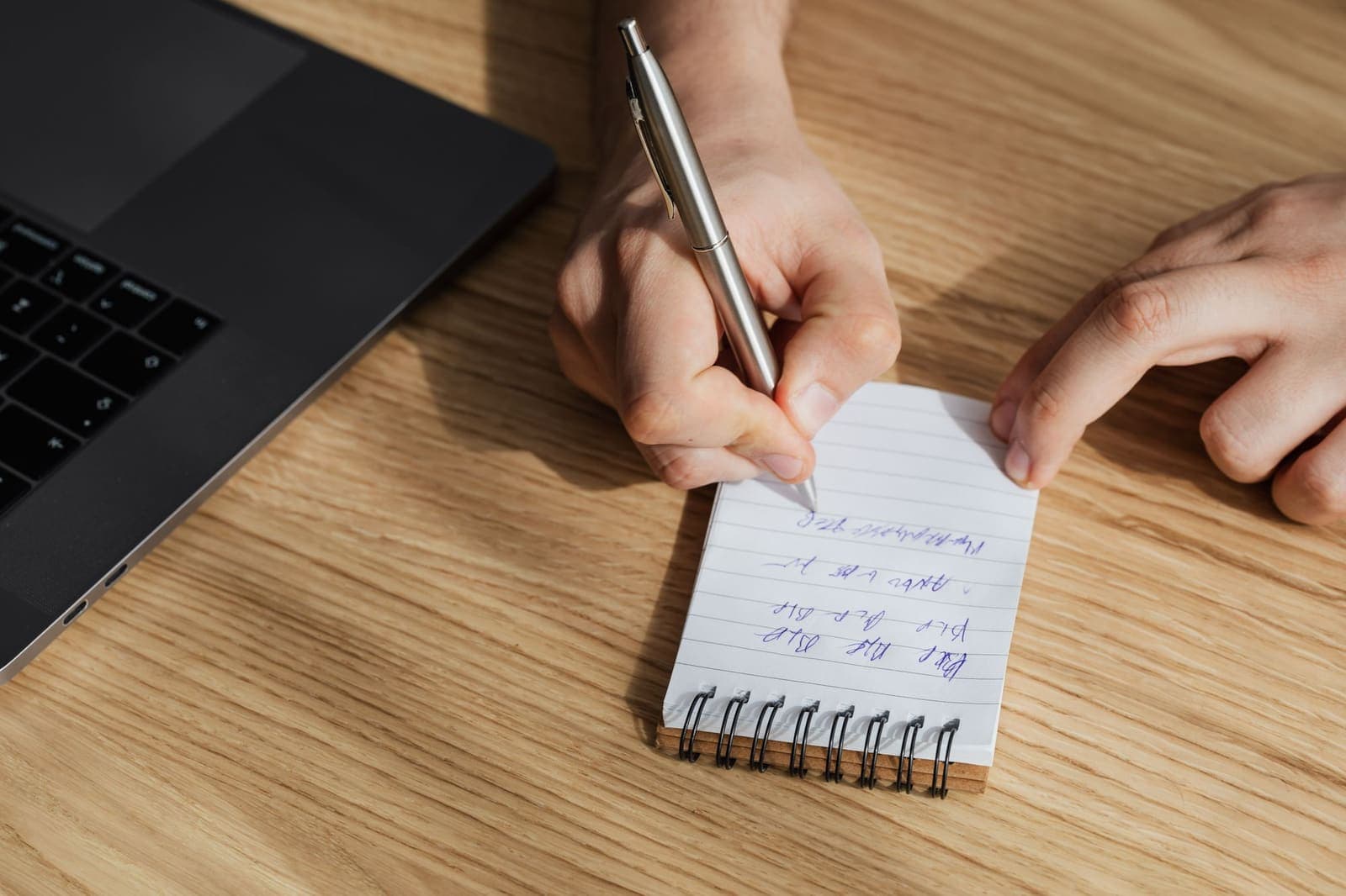 Hands writing notes in a notebook beside an open laptop