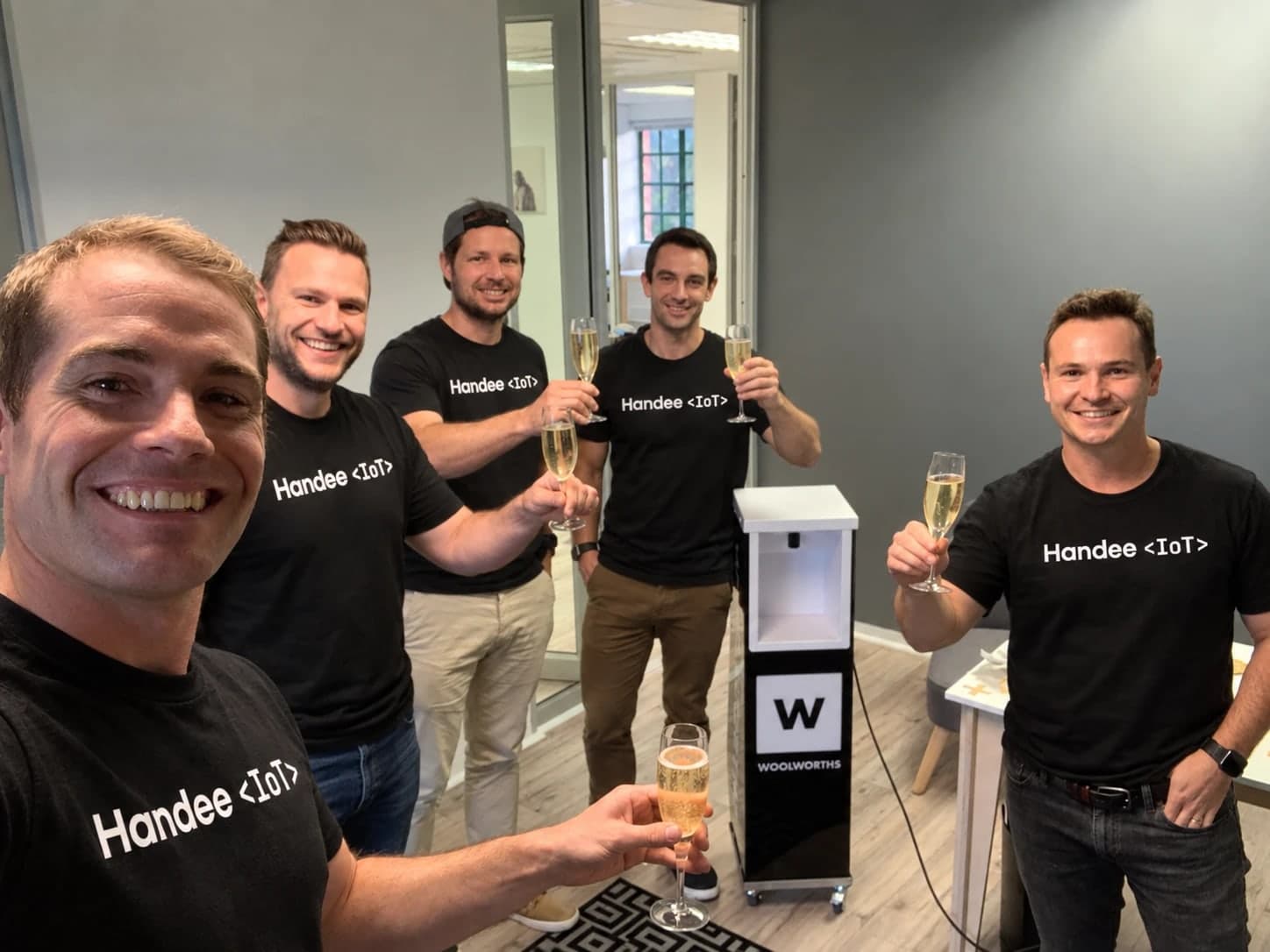 The Handee team in Handee <IoT> t-shirts, raising a glass beside the Woolworths sanitiser station they built